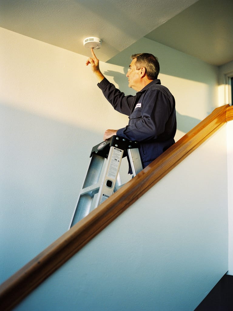A building manager on a step ladder tests a ceiling smoke detector in a stairwell.